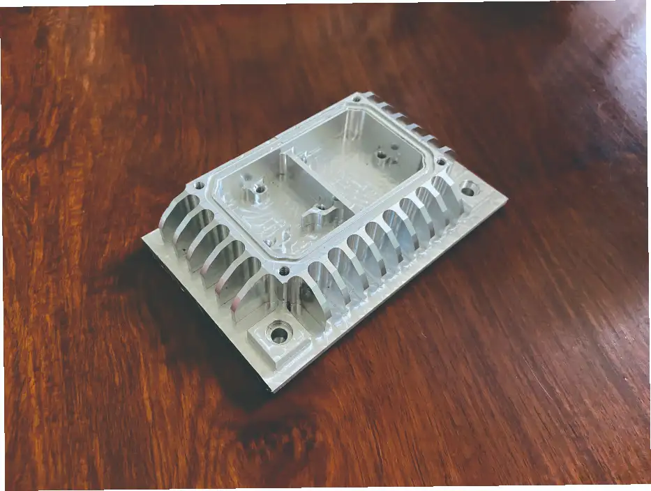 An aluminum part processed by the V-series stereo milling machine is placed on a wooden table. The part has a complex cavity, cooling fins and mounting holes, demonstrating the precision milling capabilities of the machining center.