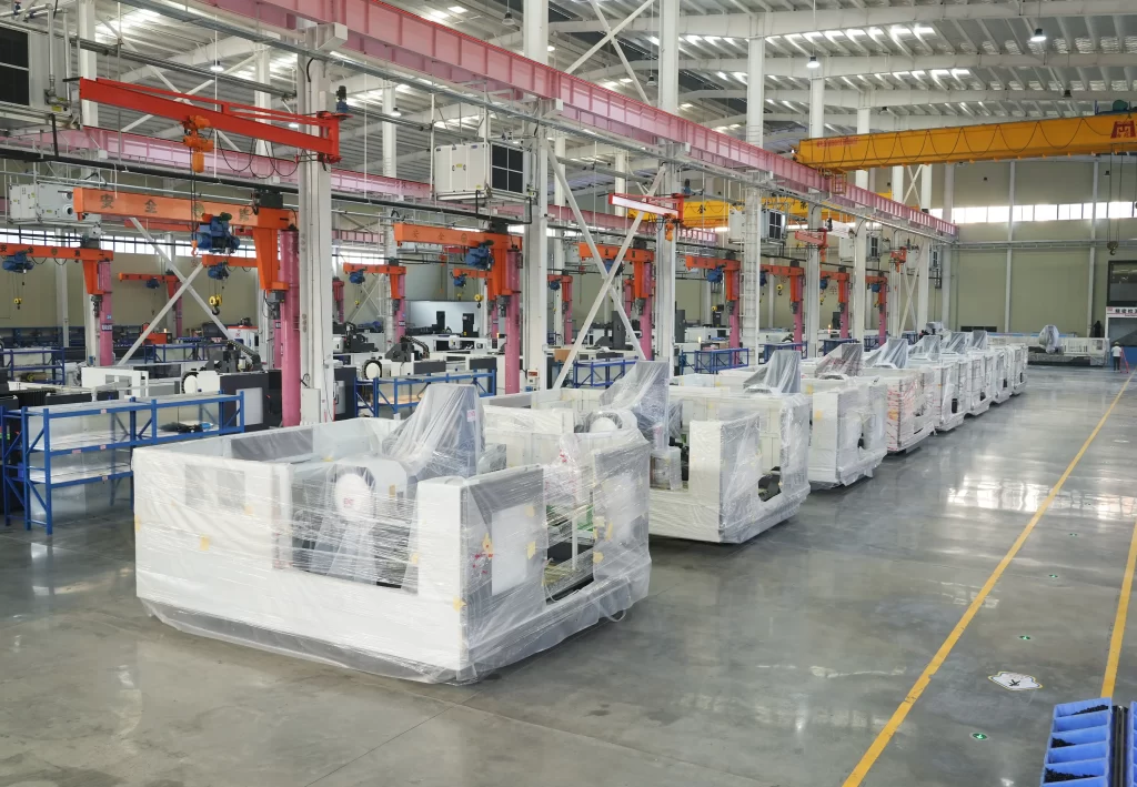 Rows of packaged CNC machines stored in an industrial factory warehouse, with overhead cranes and organized inventory layout