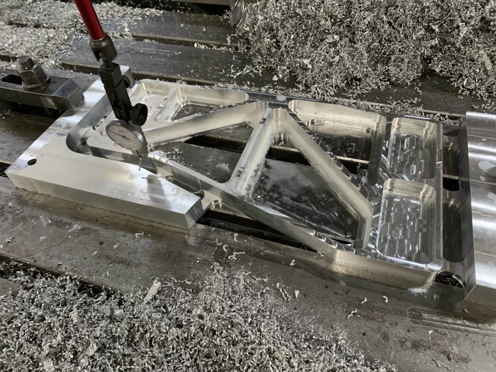 A vertical machining center mills an aluminum part, with metal chips scattered around. A red - handled tool shapes the component’s frame.
