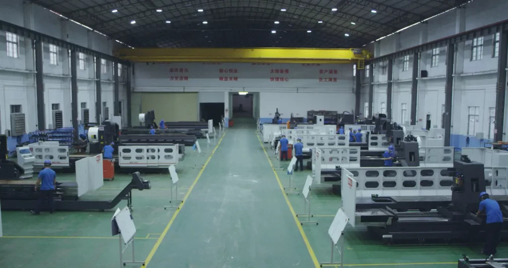 In a large manufacturing workshop, rows of CNC machines line a yellow aisle. Workers operate the machines, with banners and overhead cranes in the distance, illustrating large-scale production.