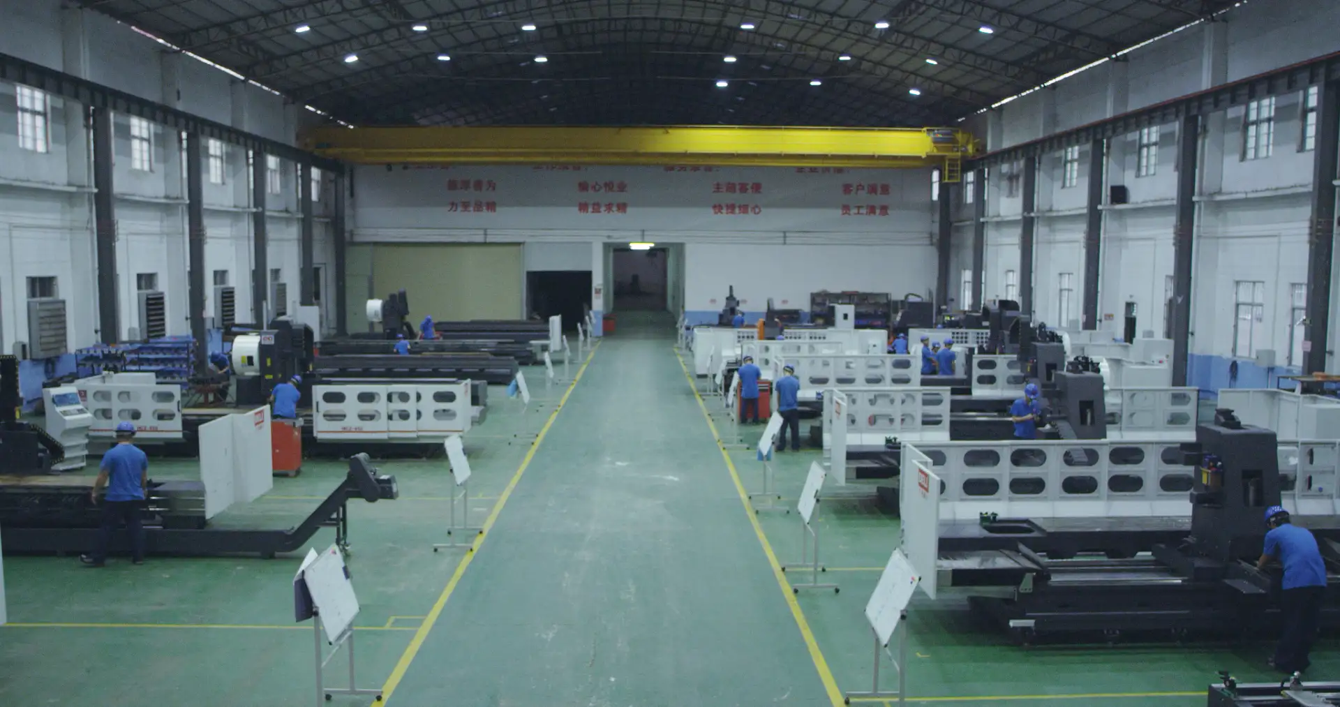 In a large manufacturing workshop, rows of CNC machines line a yellow aisle. Workers operate the machines, with banners and overhead cranes in the distance, illustrating large-scale production.