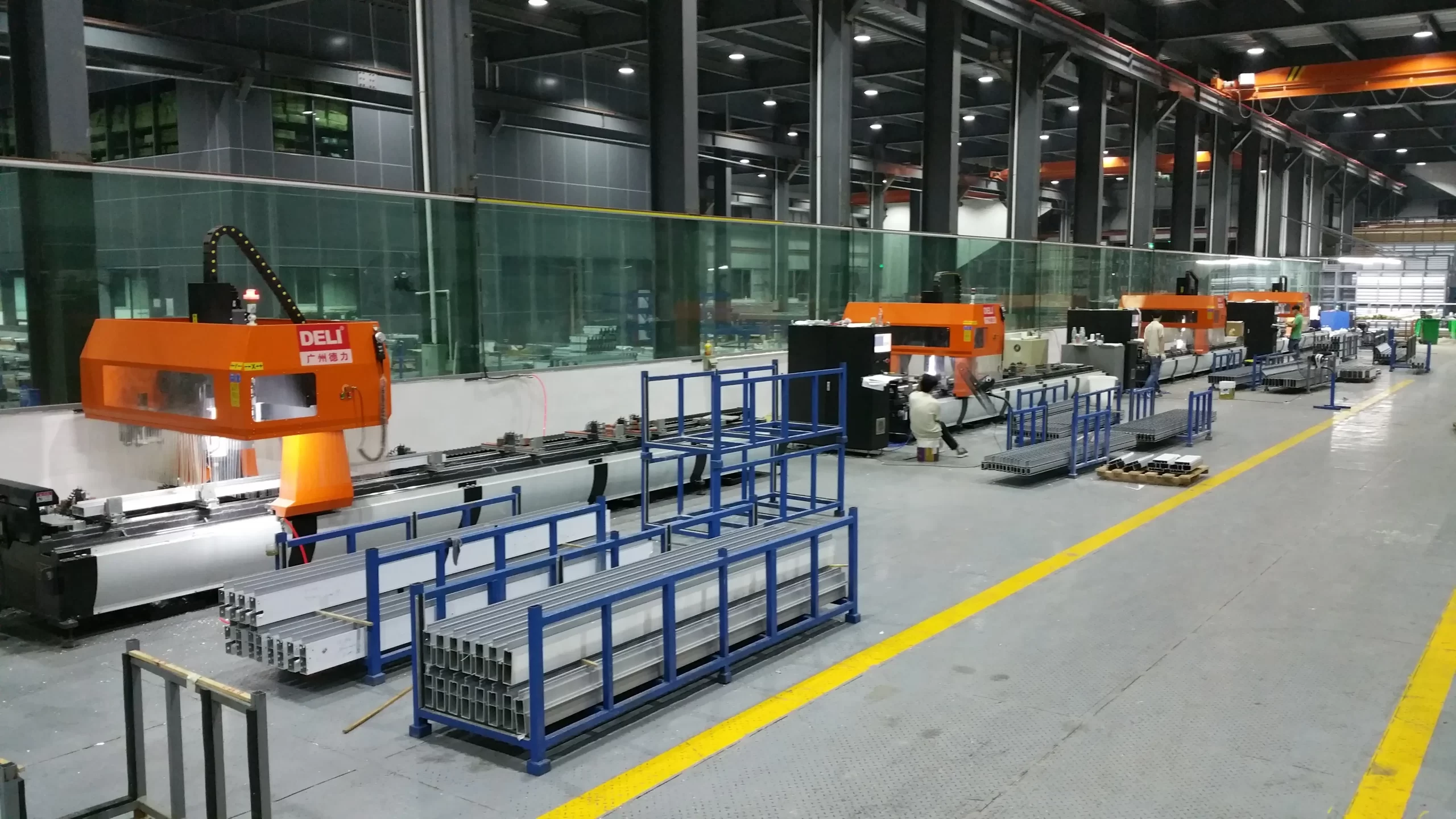 A spacious industrial factory with large CNC machinery and aluminum profiles on blue racks, plus an orange robotic arm and glass partitions.