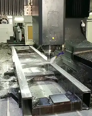 A large CNC milling machine cutting a substantial metal part, with coolant being sprayed onto the cutting area and chips covering the machine and workpiece.