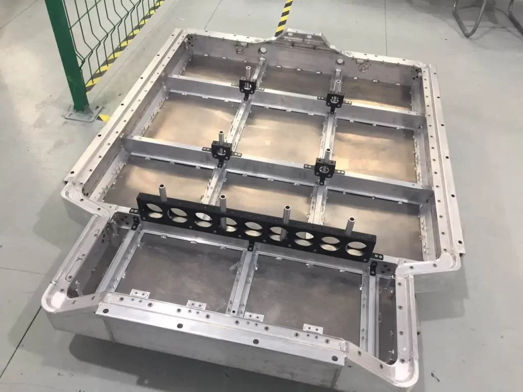 A large aluminum battery tray component, machined with precision, featuring multiple slots, mounting points, and structural reinforcements, placed in a clean industrial environment.
