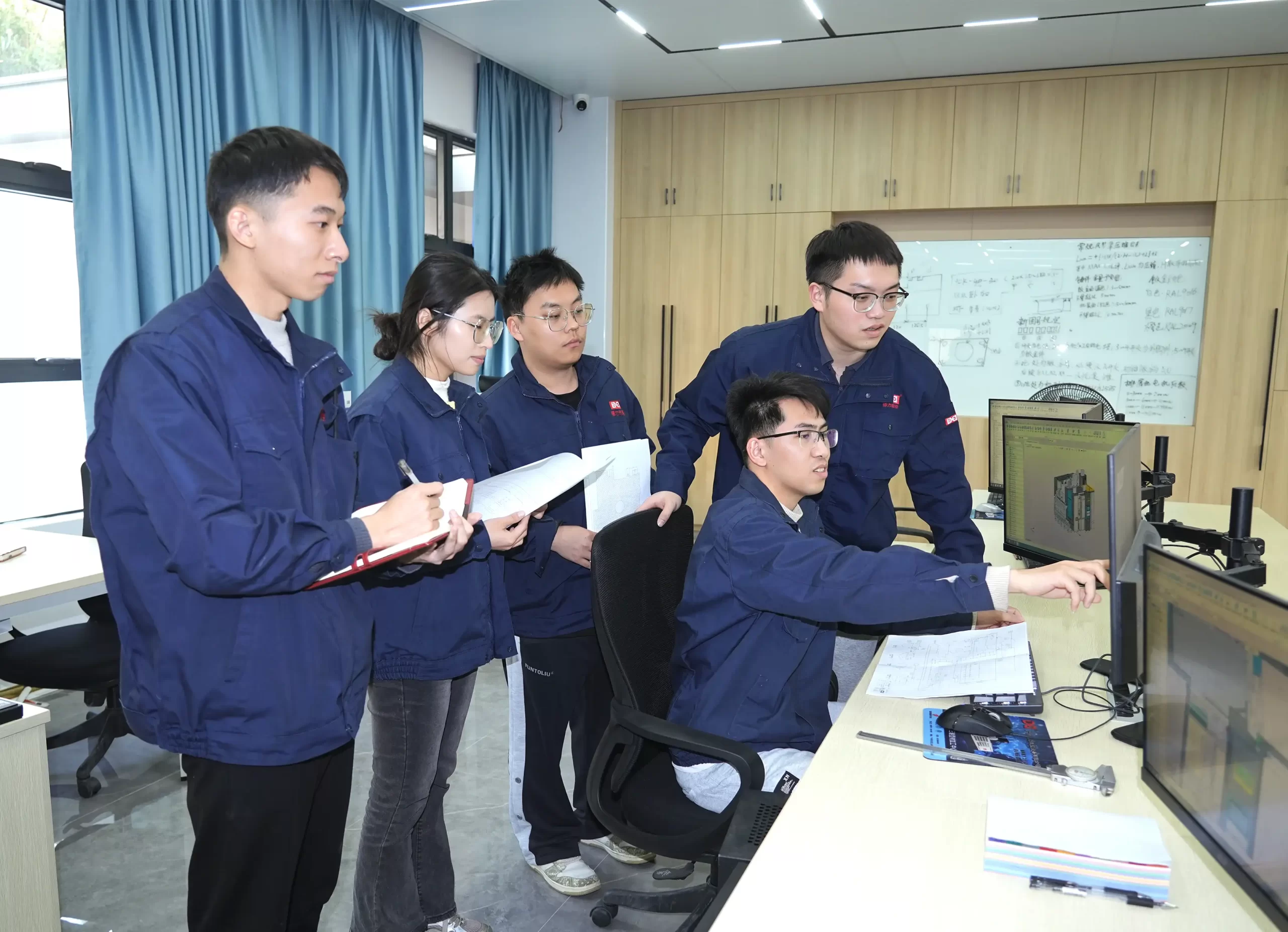 A group of CNC operators in blue work uniforms collaborate in an office, focusing on computer-aided design (CAD) models on monitors, with documents and a whiteboard with technical diagrams in the background.