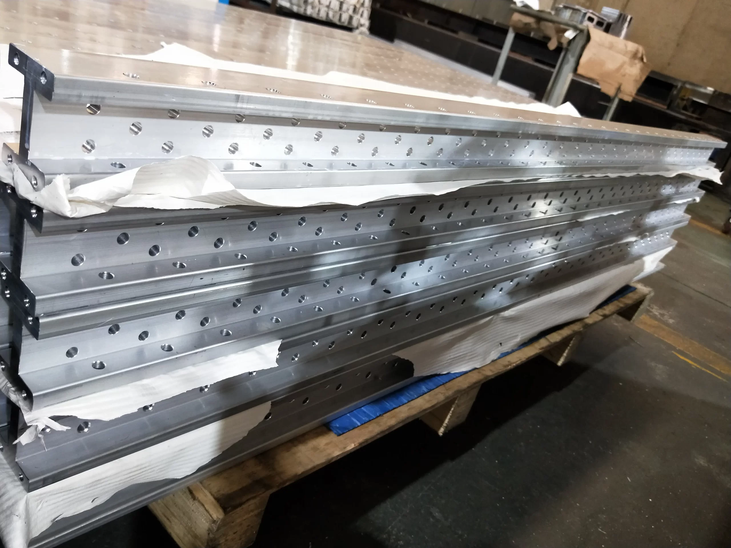 Stack of precision-machined aluminum components with evenly spaced holes, wrapped in protective paper, placed on a wooden pallet in an industrial manufacturing workshop setting.